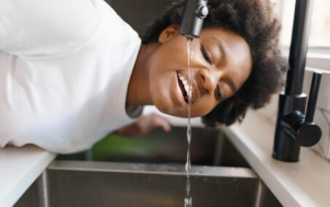 A black woman drinking water directly from a kitchen faucet